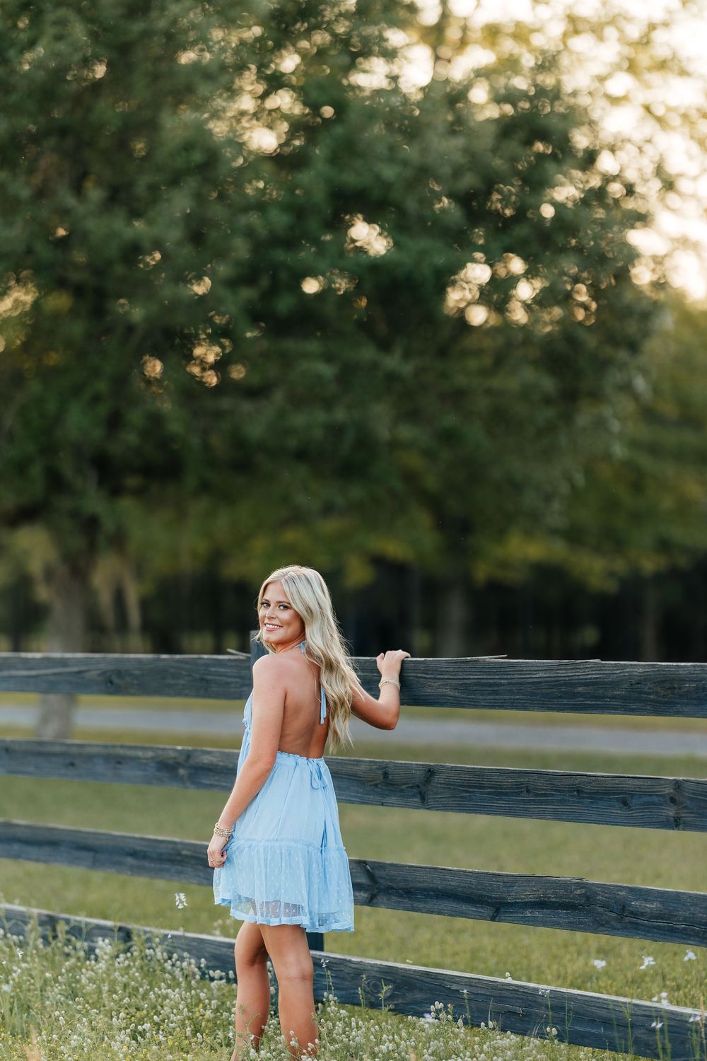 Woman in light blue summer dress poses near rustic wooden fence at sunset in grassy field.