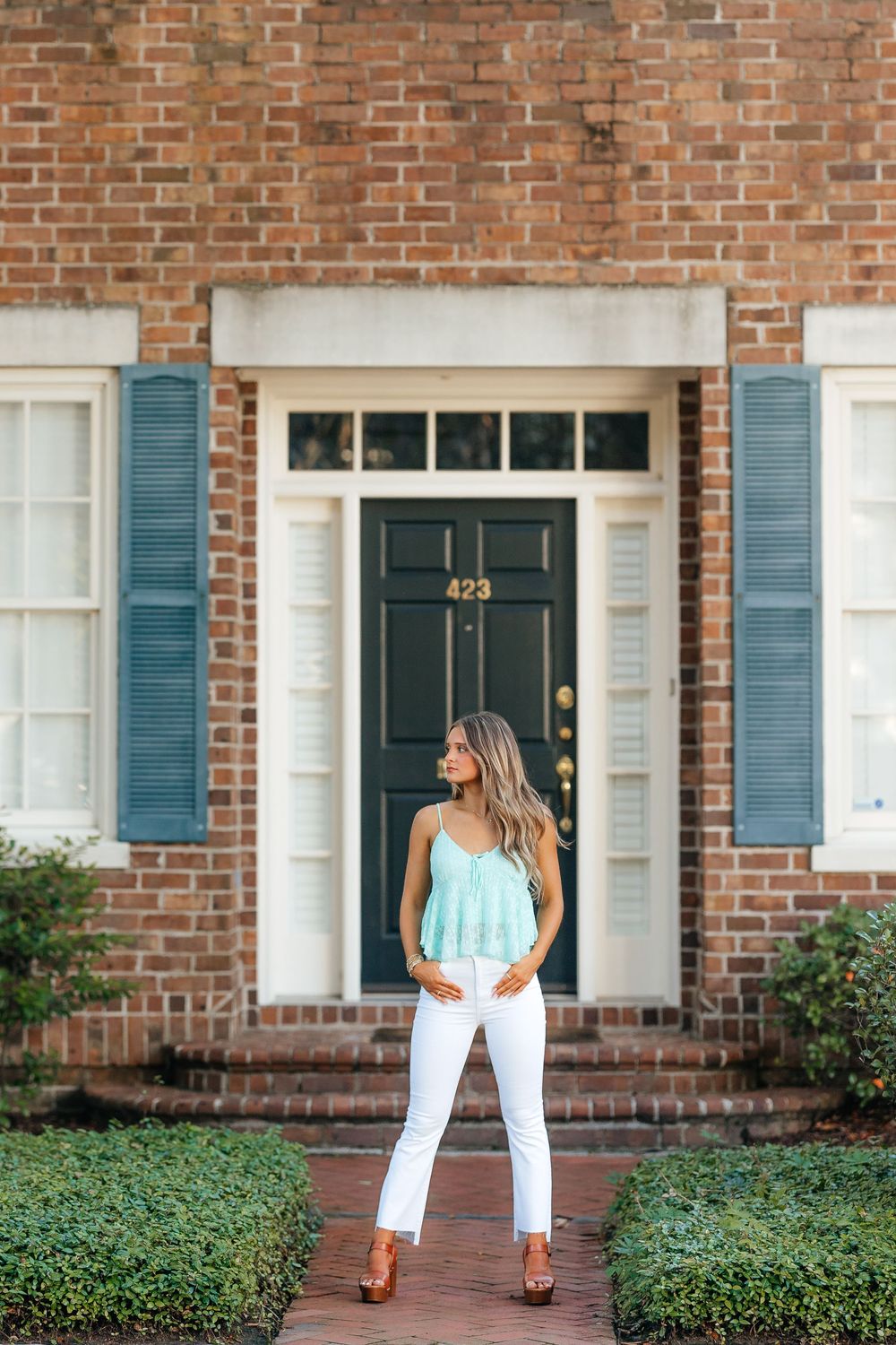 Person in white pants and blue top stands in front of traditional brick home with blue shutters and dark front door.