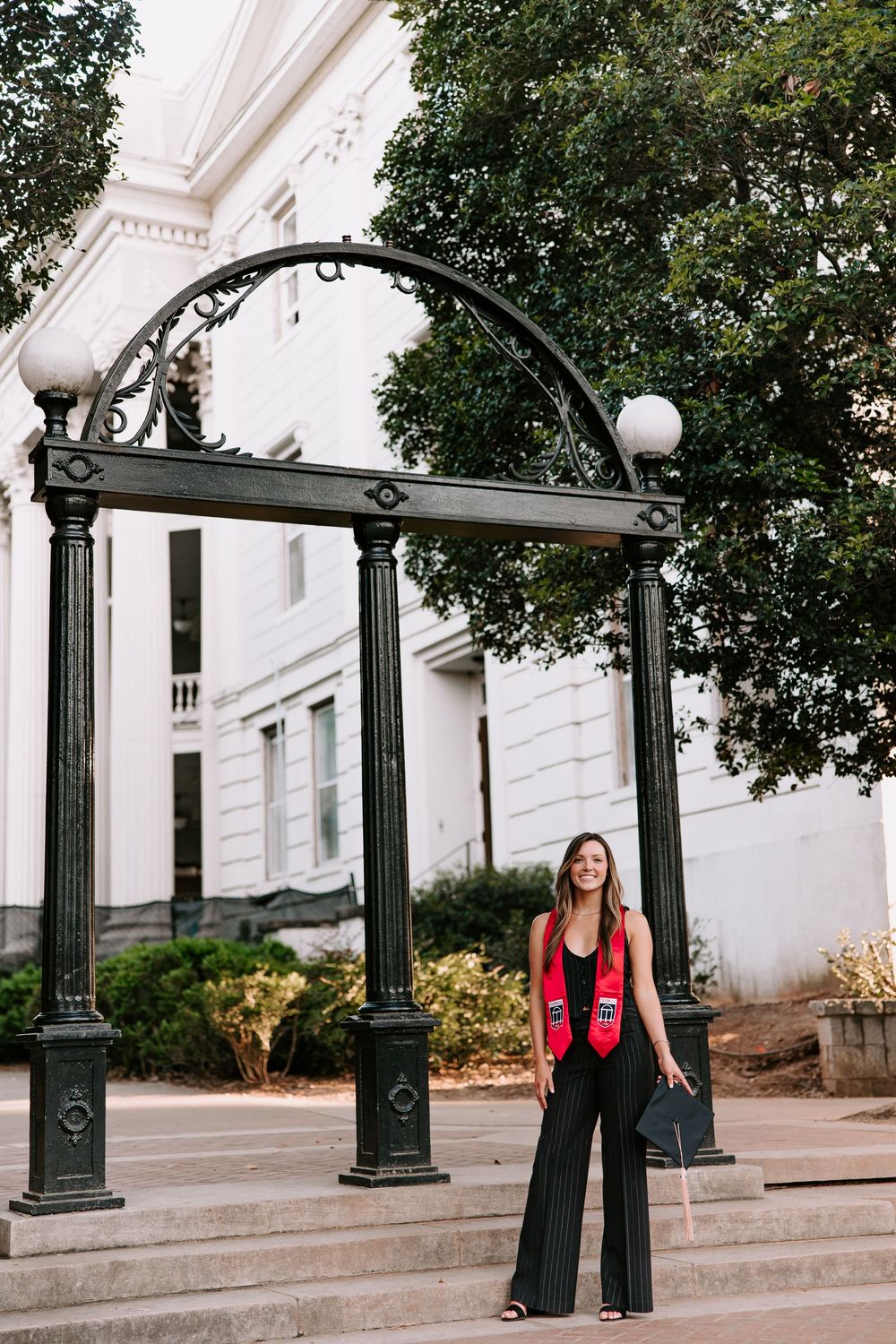 A stylish figure poses beneath an ornate black iron arch entrance with white columns and trees in the background.