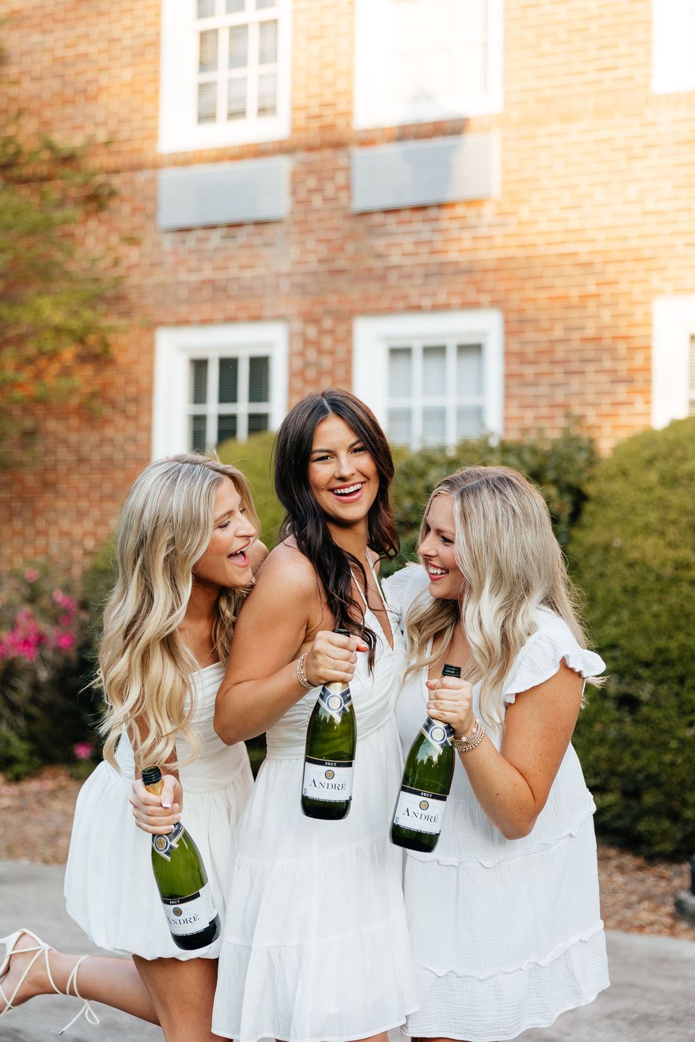Friends in matching white outfits celebrate with champagne bottles outside a brick building.