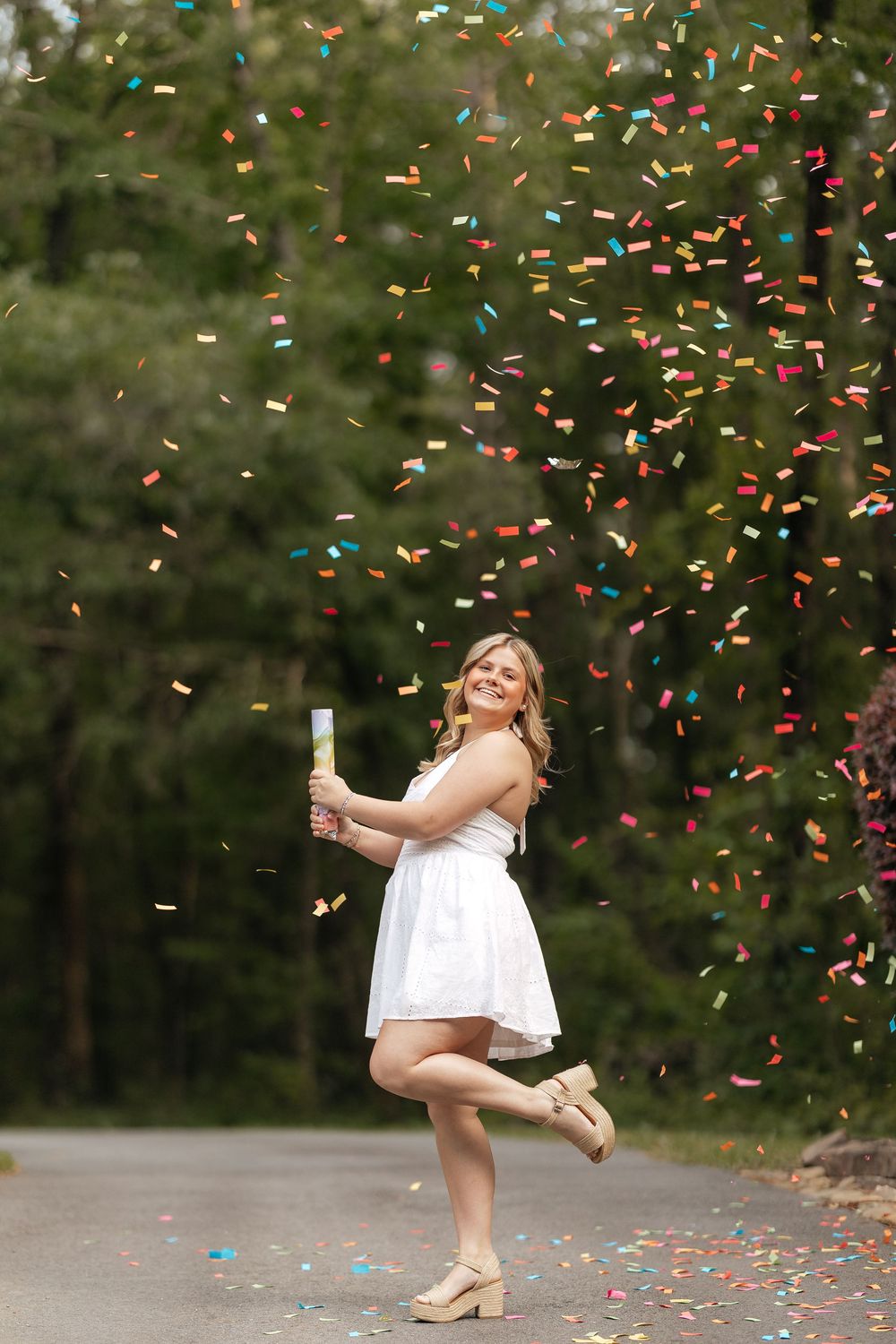 A joyful young woman in a white dress celebrates outdoors with colorful confetti raining down around her.