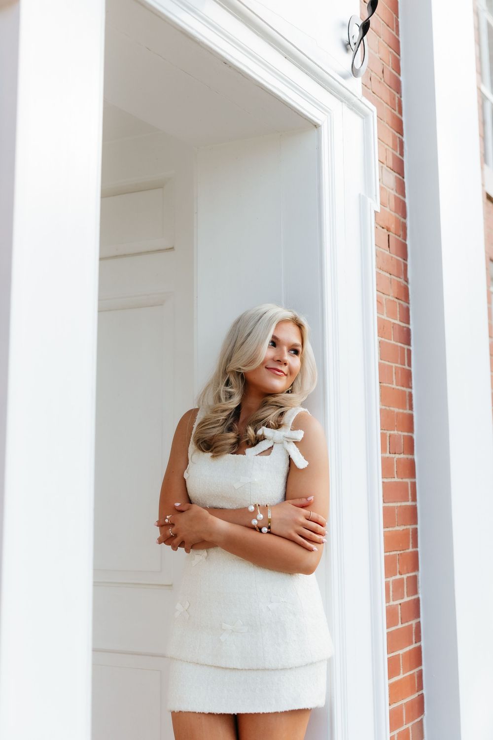 Blonde woman in white mini dress with floral detail poses gracefully in a white doorway beside a brick wall.
