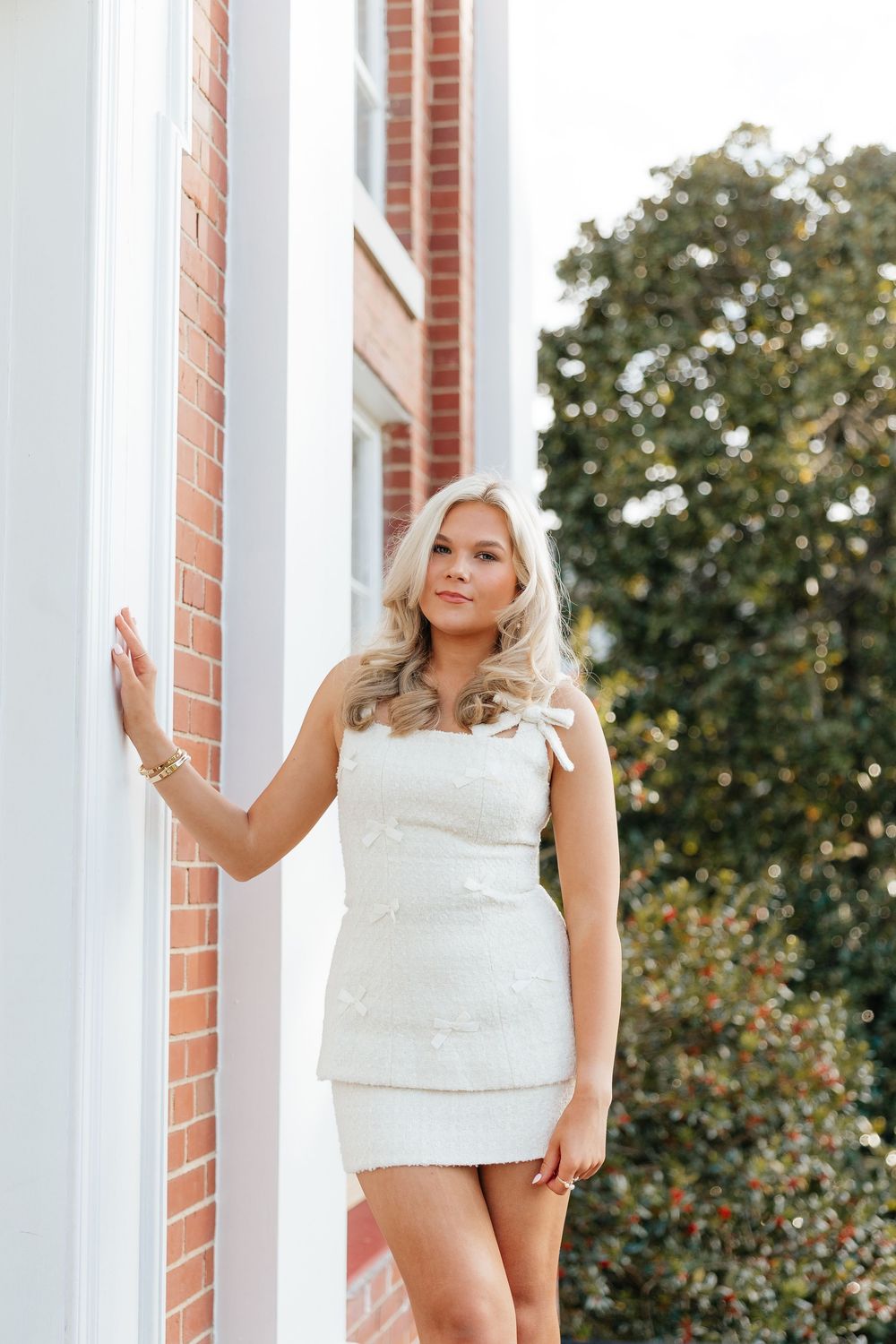 Blonde woman in white mini dress posing against brick column outdoors with green trees in background.