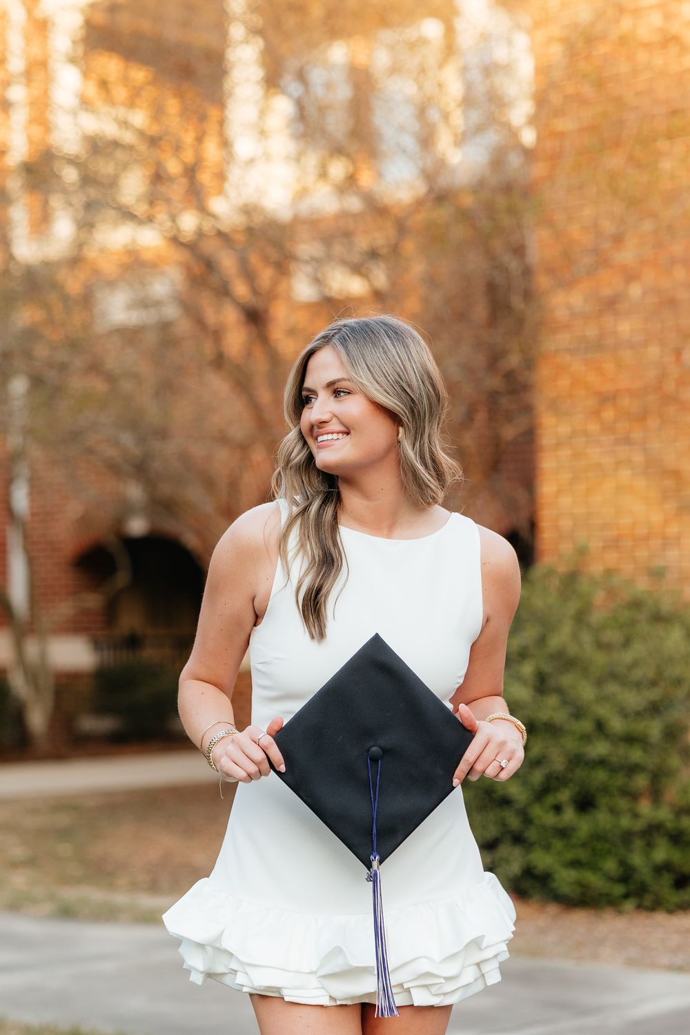 Smiling young woman in white dress holding graduation cap outdoors near brick building with autumn foliage.