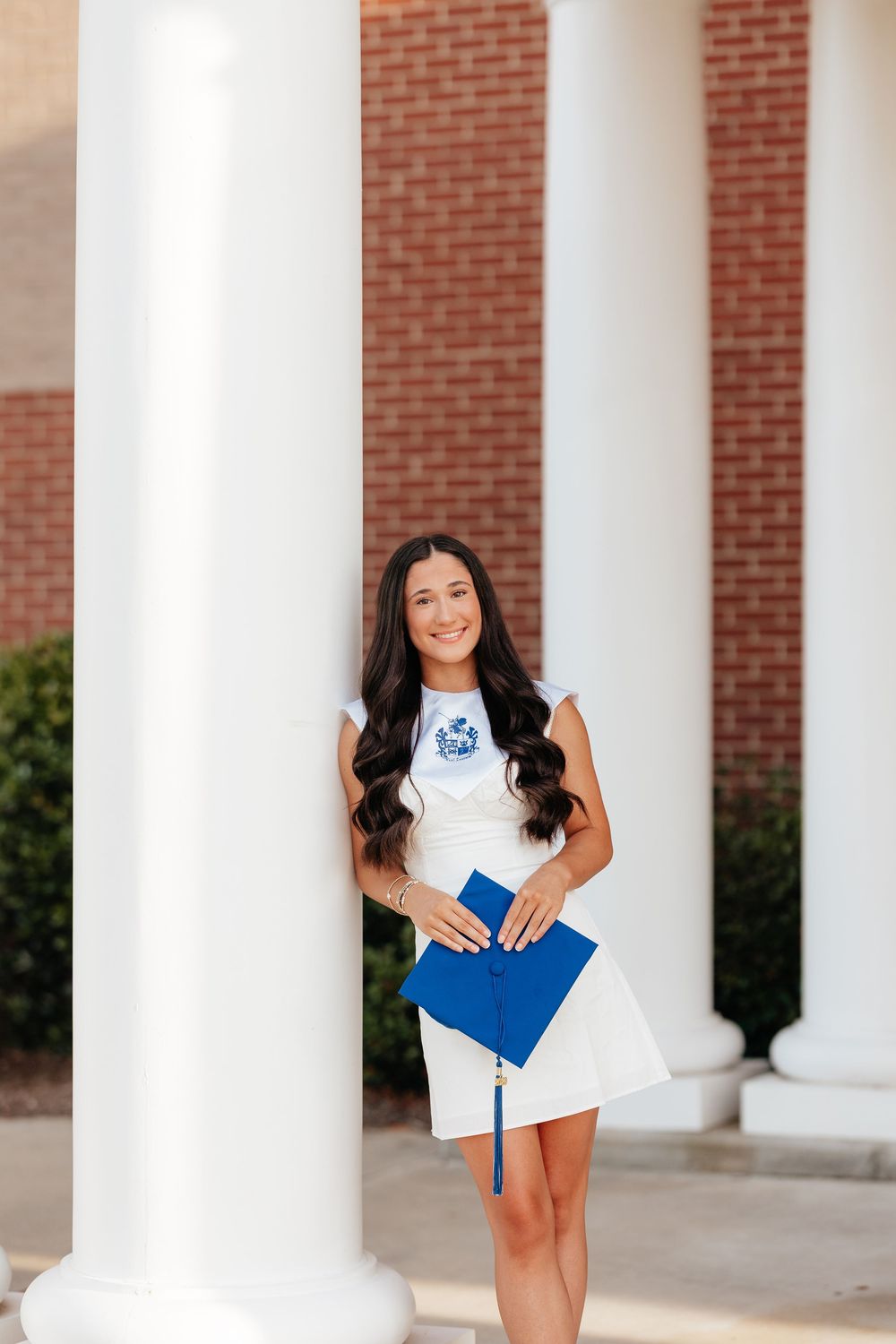 Young woman in white dress holding blue graduation cap poses between white columns of brick building.