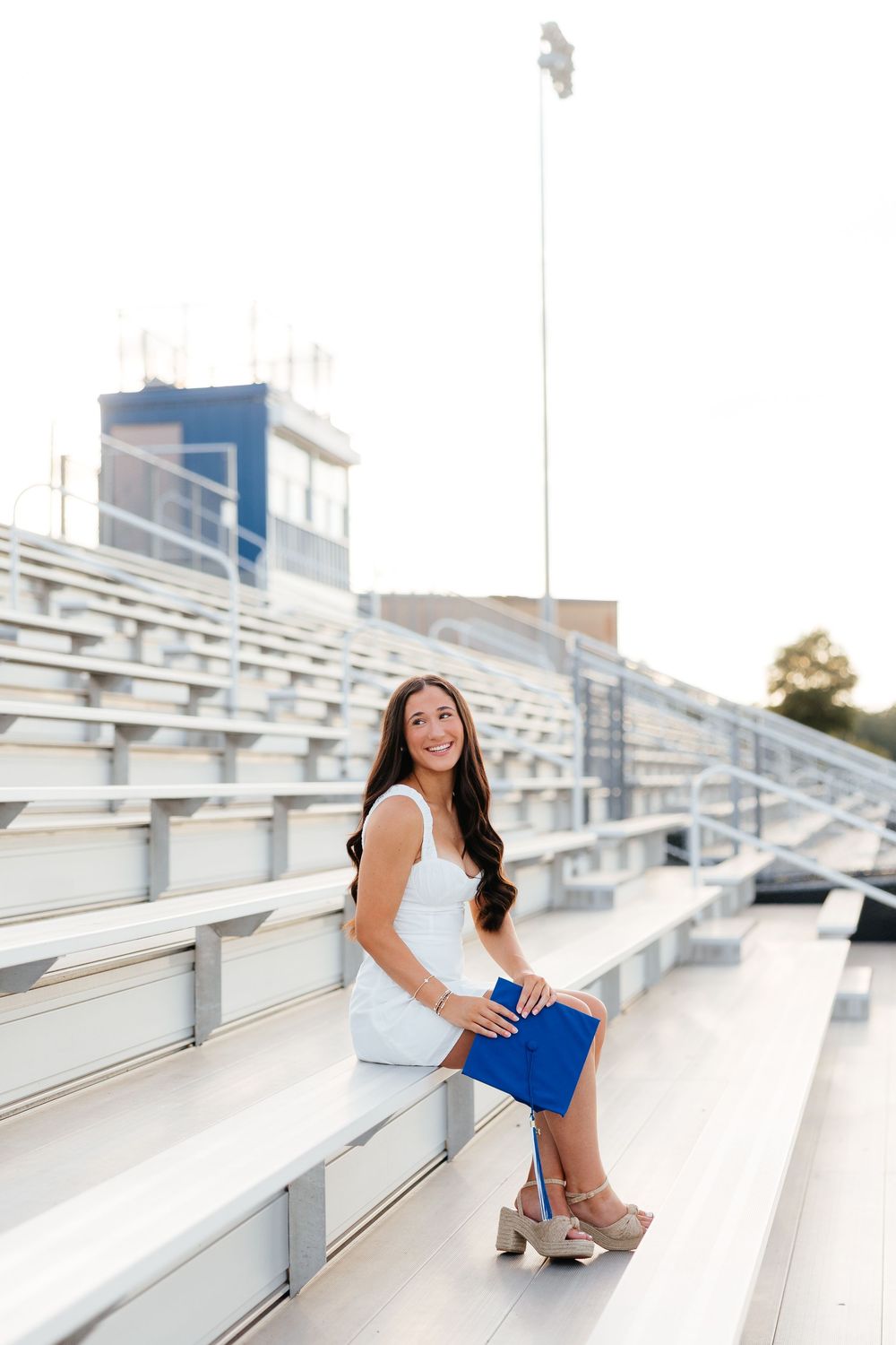 Young woman in white dress holding blue diploma sitting on stadium bleachers, smiling at camera during graduation photo shoot.