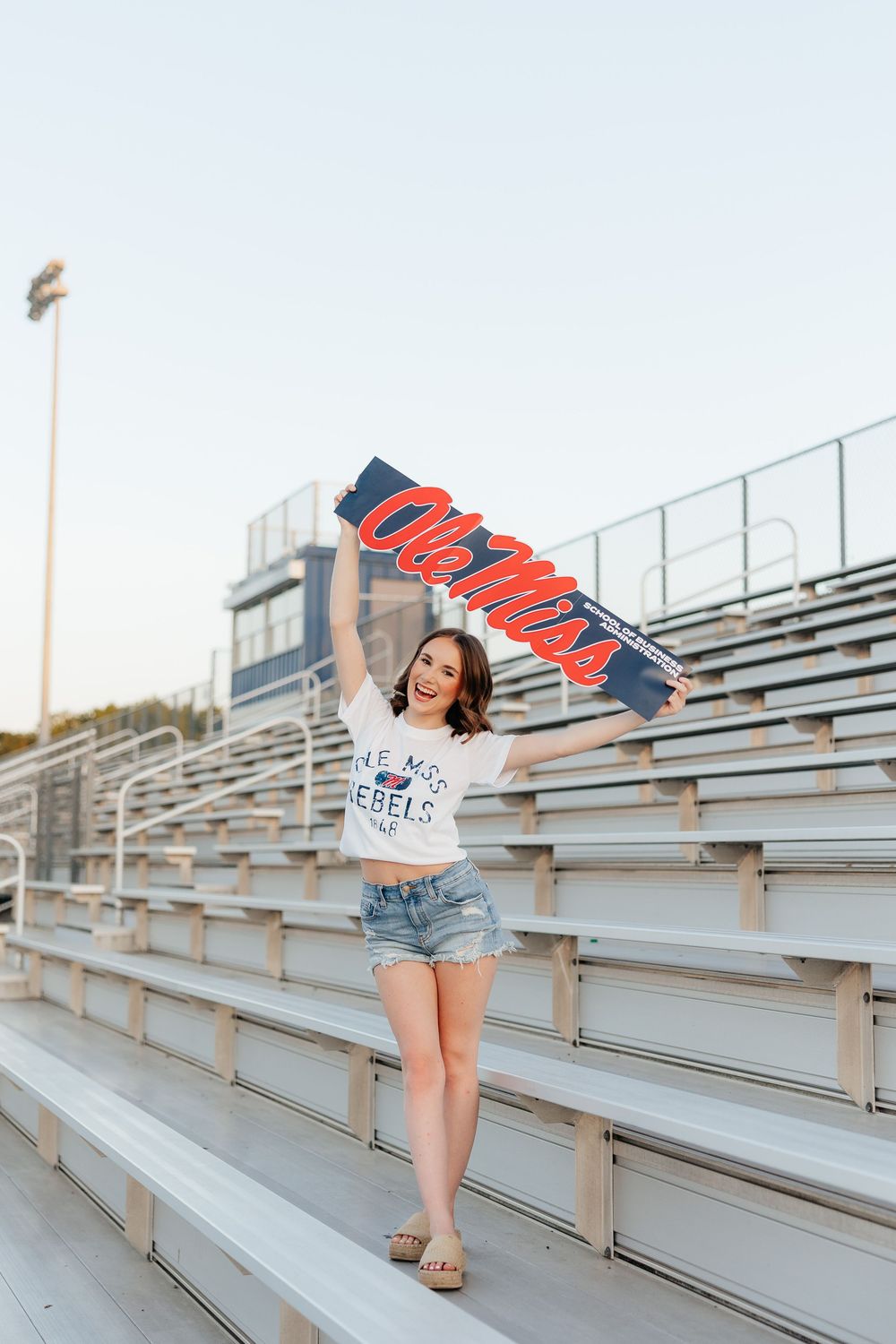 Smiling young woman holding Ole Miss Rebels flag in stadium bleachers wearing white tee and denim shorts.