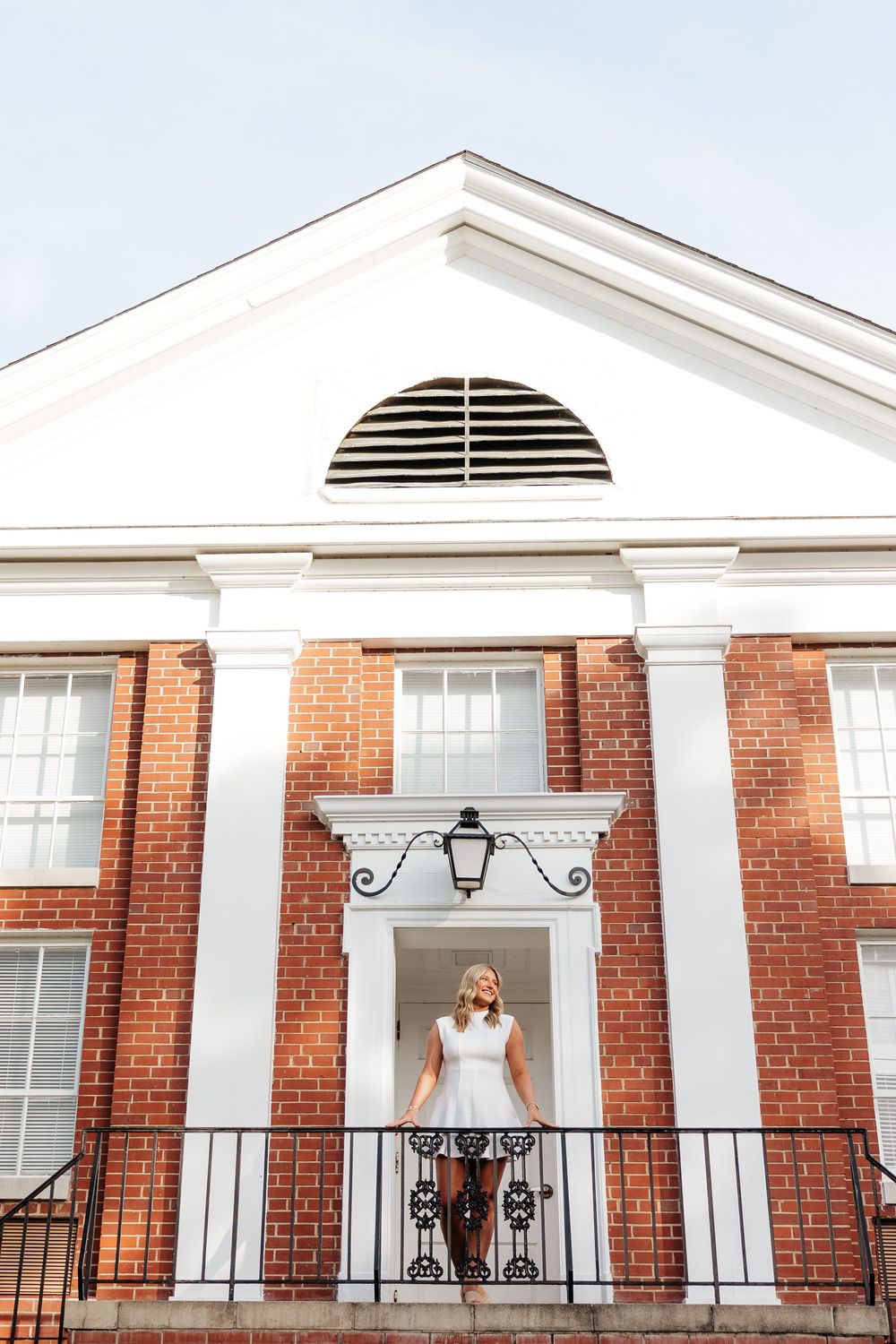 Bride in white dress standing on balcony of elegant red brick building with white columns and pediment.