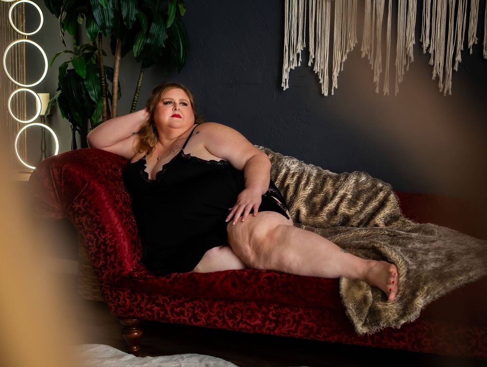 Plus-size woman in black dress reclines elegantly on a red velvet chaise lounge with fur throw in a moody studio.