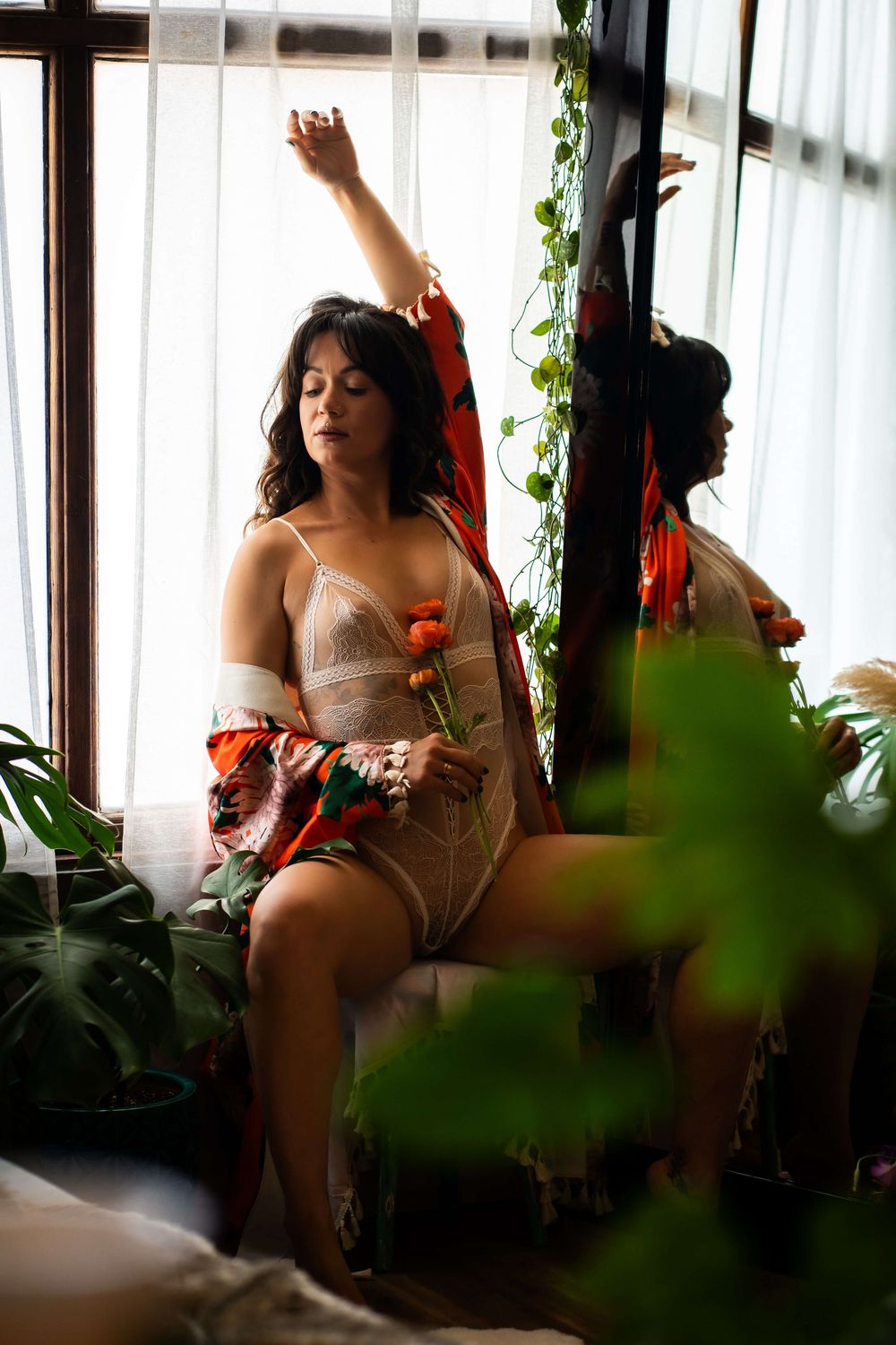 Woman in lace bodysuit holding orange flowers, seated by window with plants and mirror reflection.