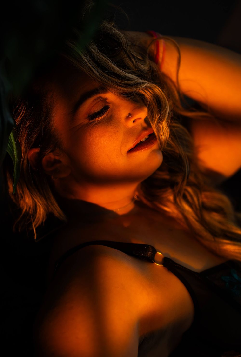 Woman with wavy blonde hair posed in dramatic warm orange lighting against a dark background.