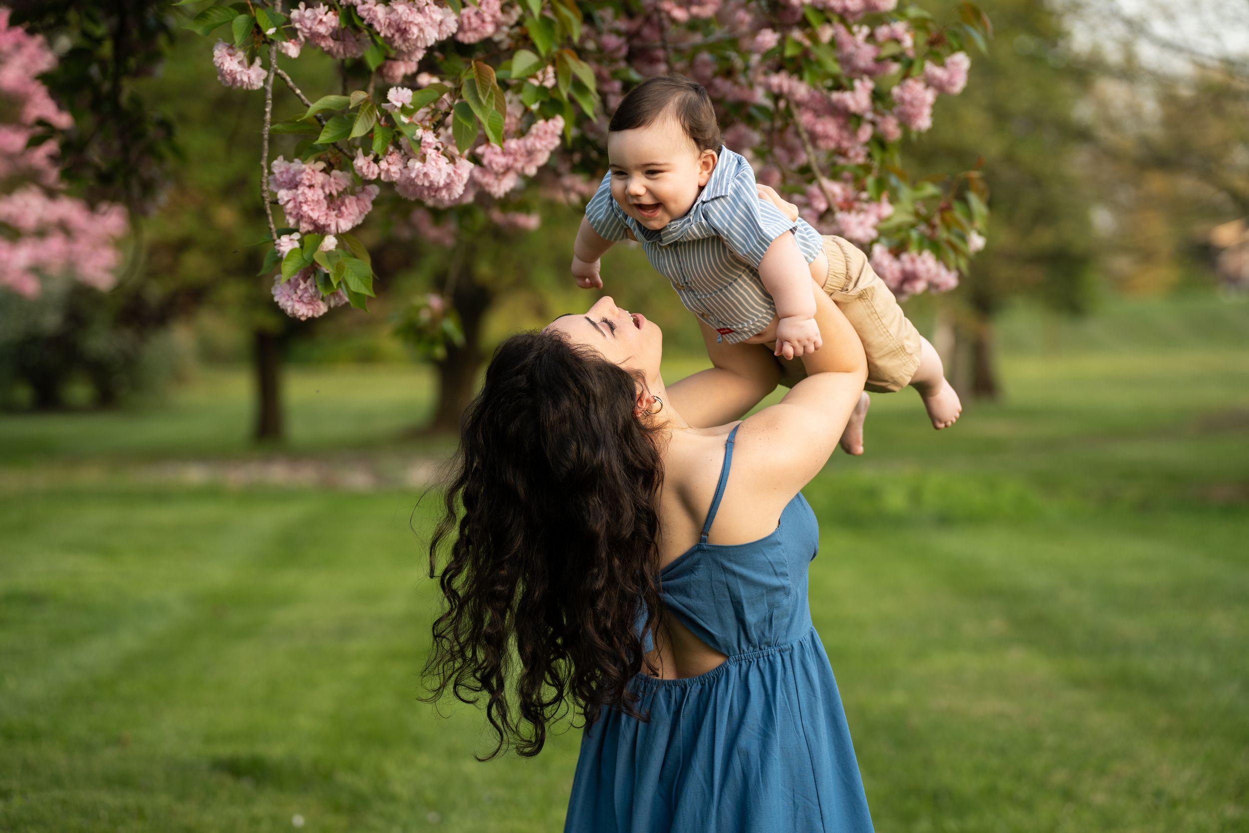 Mother in flowing blue dress lifts baby under blooming cherry blossom tree in spring park.