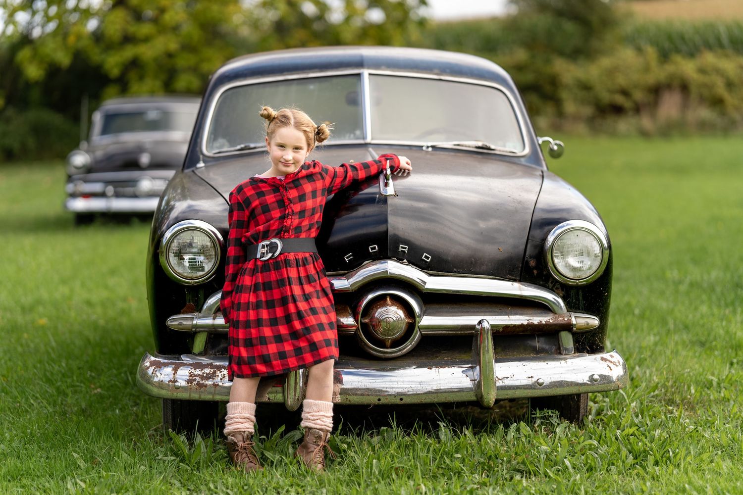 A child in a red plaid dress poses playfully in front of a vintage black car.