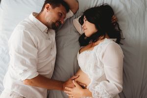 A couple wearing white shirts lies together on white bedding while sharing an intimate moment during a maternity photo session.