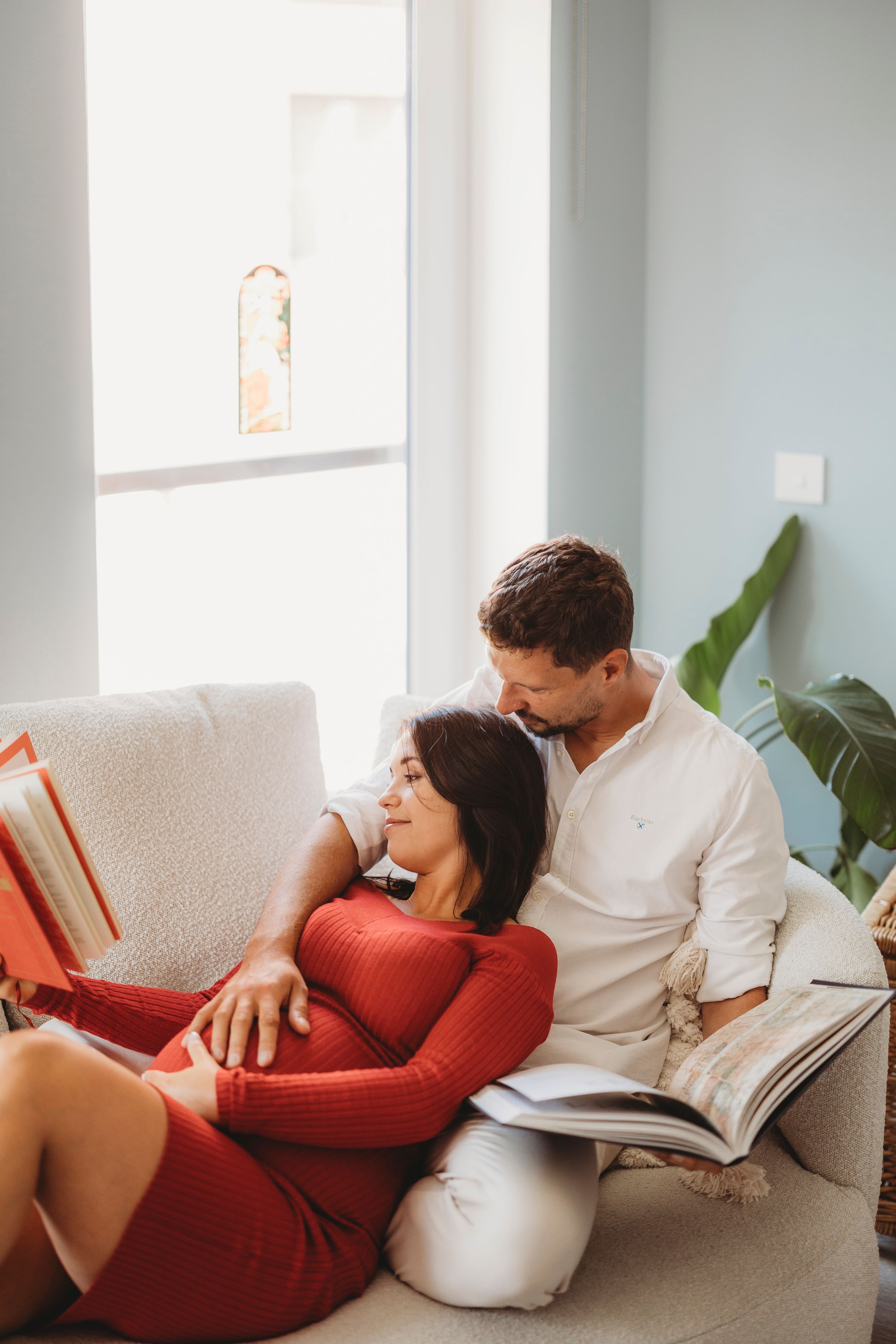 Couple relaxing together on couch reading books in bright sunlit room with houseplant by window.