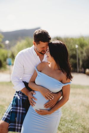 A romantic outdoor maternity photoshoot shows a couple embracing in a sunlit field with mountains in the background.