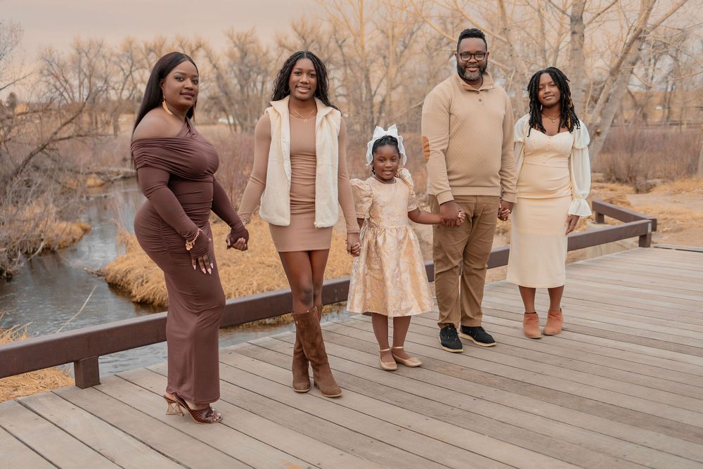 A family of five poses on a wooden bridge in a scenic autumn outdoor setting, dressed in coordinated neutral tones.