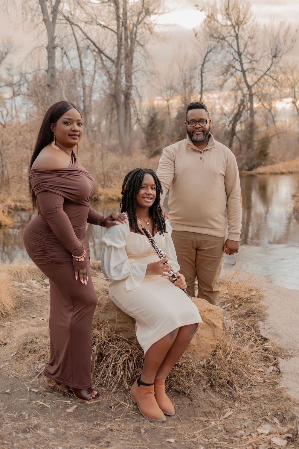 A family of three poses outdoors near a pond, dressed in elegant earth-tone outfits, surrounded by autumn trees.