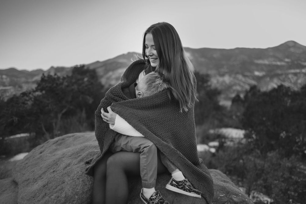 Black and white photo of a laughing woman hugging a child wrapped in a blanket on a rocky hilltop.