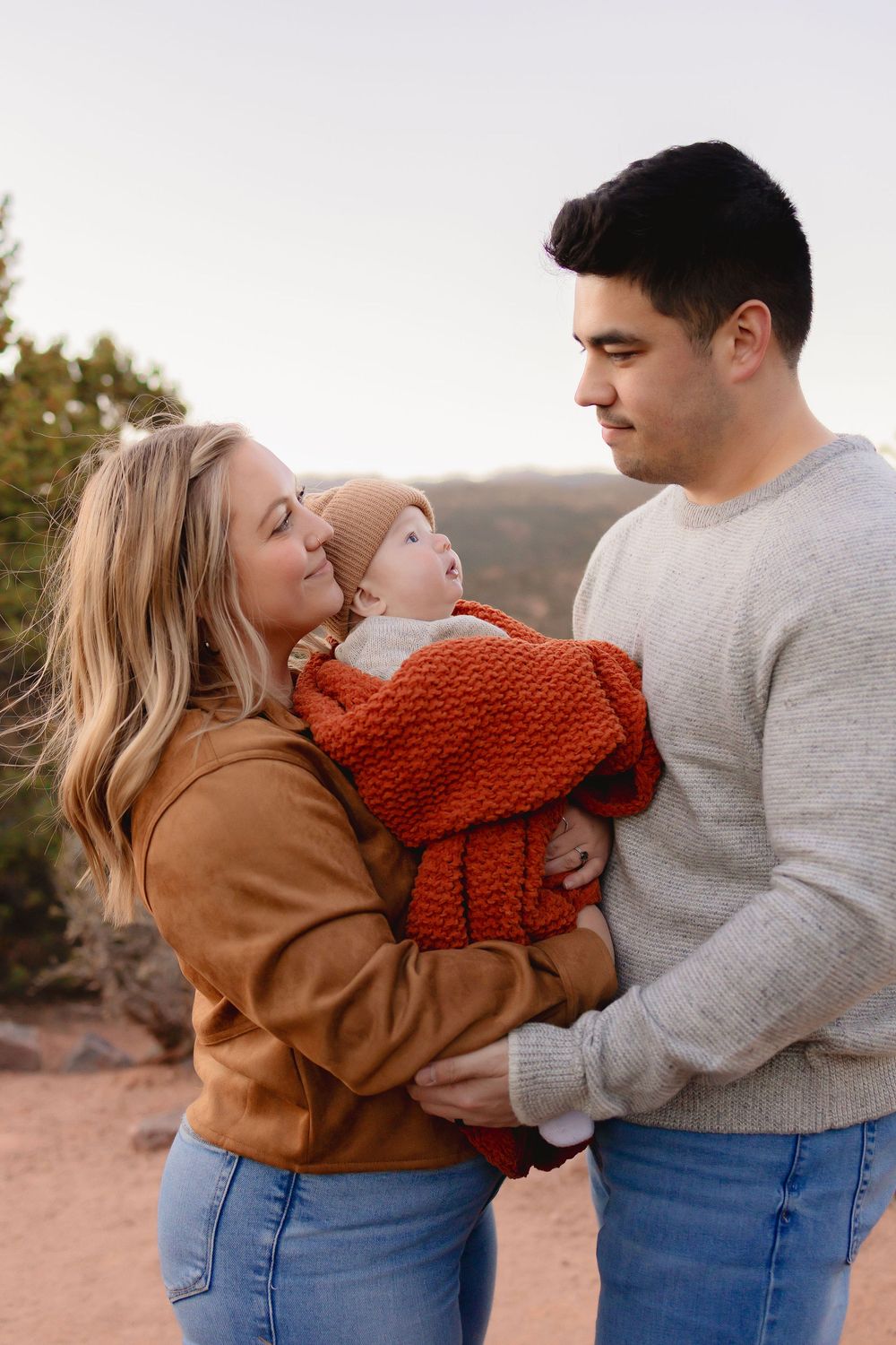 Young family with mother, father, and baby dressed in autumn colors posing outdoors in a natural landscape.