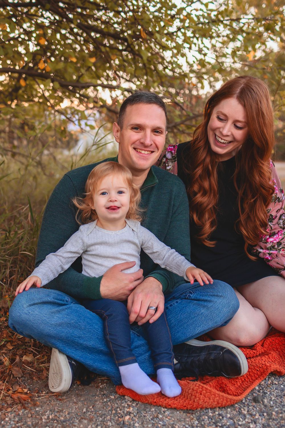 Happy family with mom, dad, and toddler sitting on orange blanket outdoors in autumn setting.