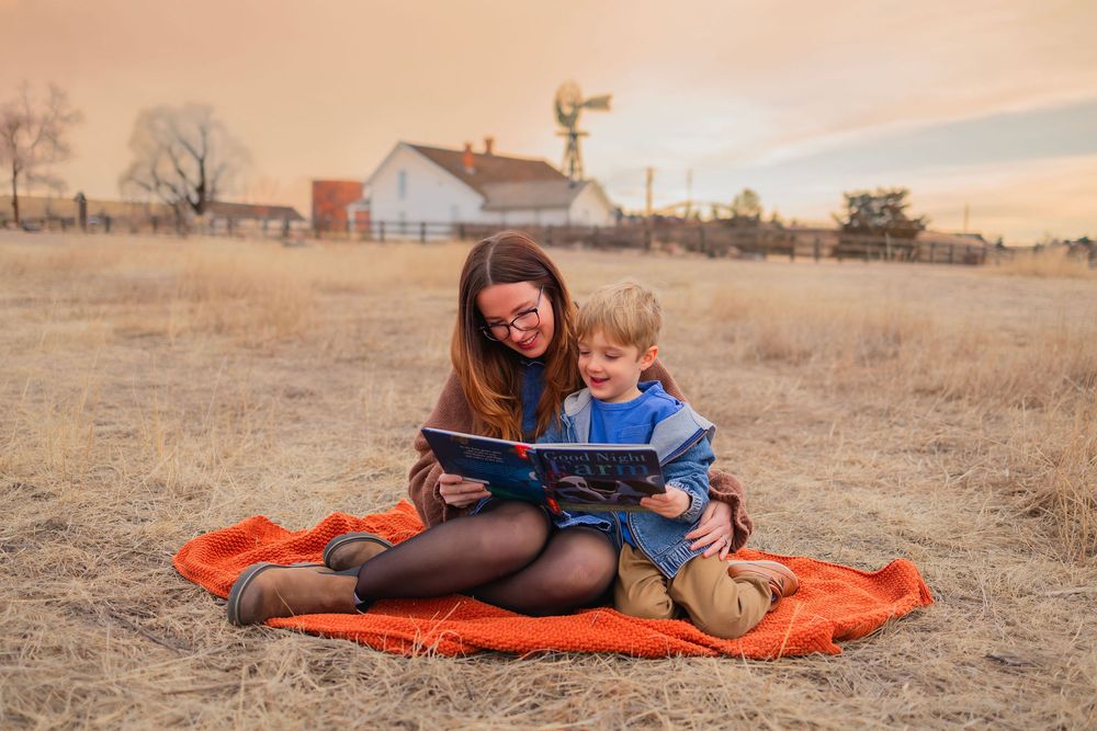 A mother and young son sit on an orange blanket in a field, reading a book together at sunset near a farmhouse.
