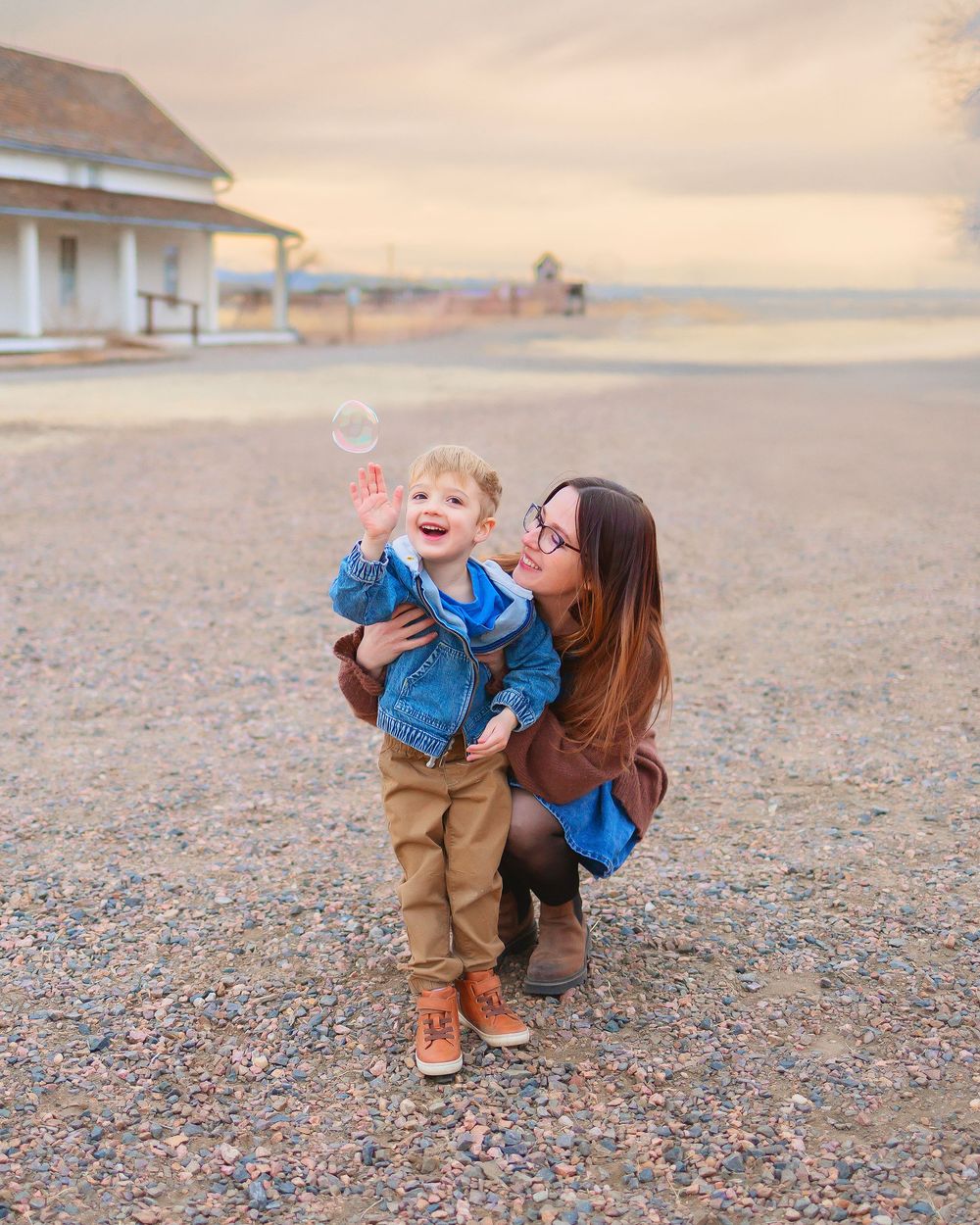 A woman crouches beside a smiling toddler reaching for a bubble outdoors at sunset.
