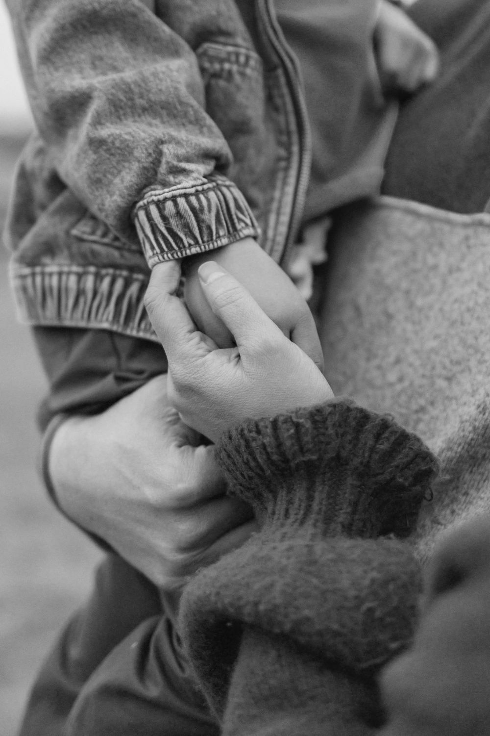 Black and white close-up of two hands holding gently, wearing knit and denim sleeves.