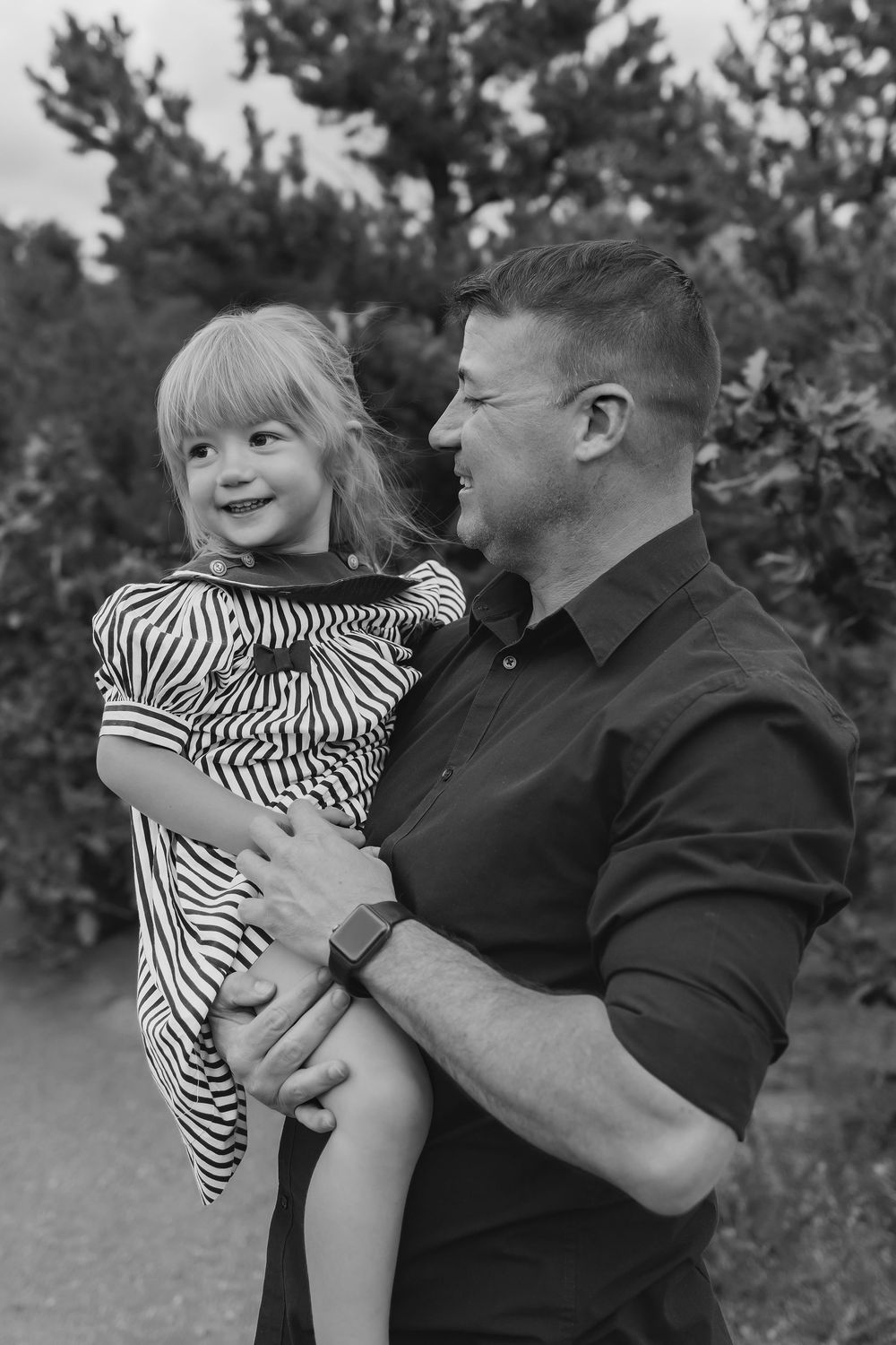 Black and white photo of a smiling man holding a young girl in a striped dress outdoors near trees.