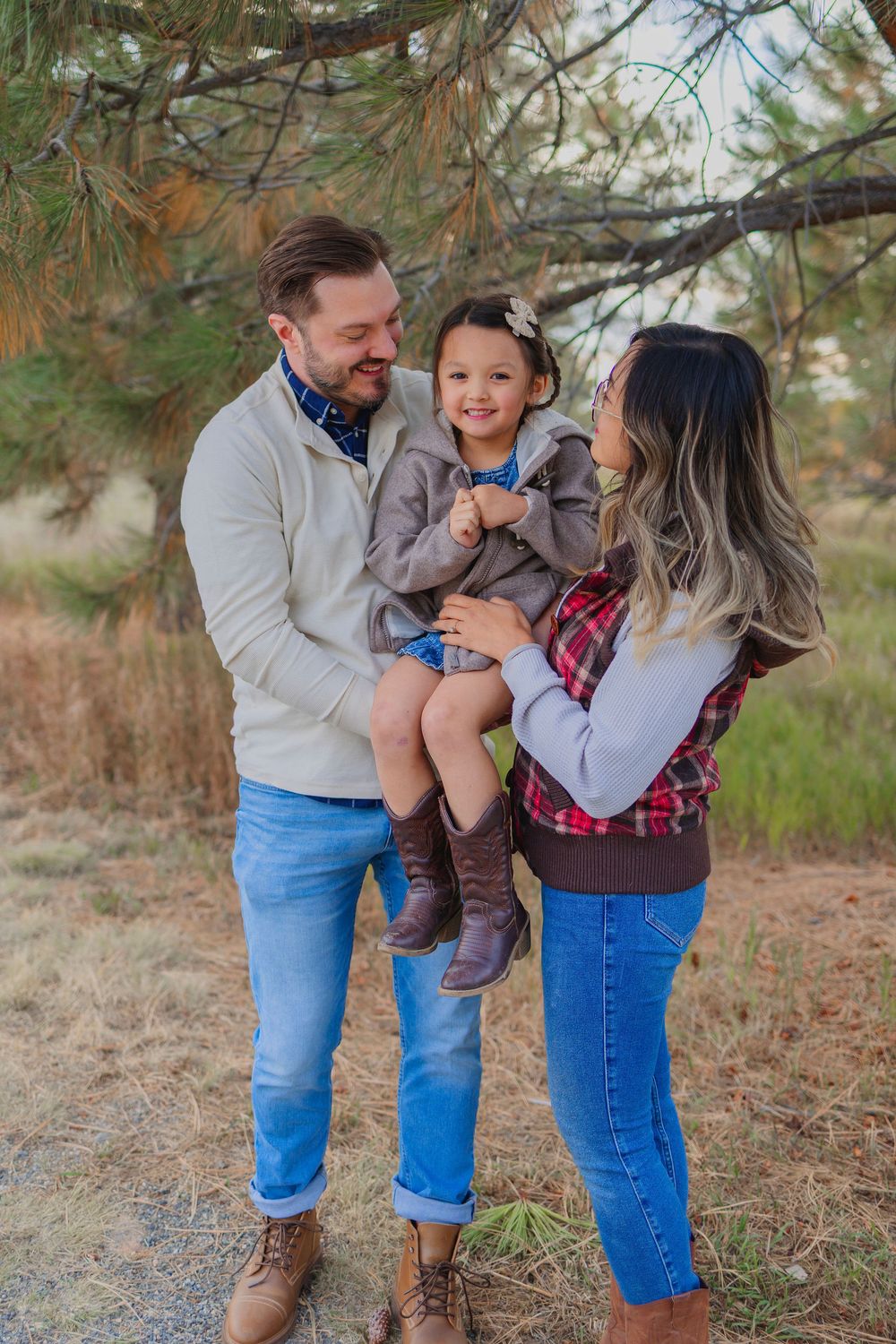 A happy family of three poses outdoors under pine trees, parents holding their young daughter in casual fall attire.