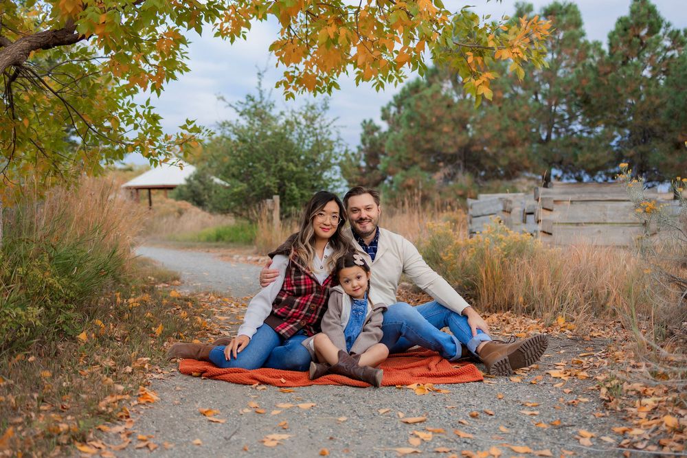 Family of three sitting on orange blanket outdoors in autumn park with colorful foliage.