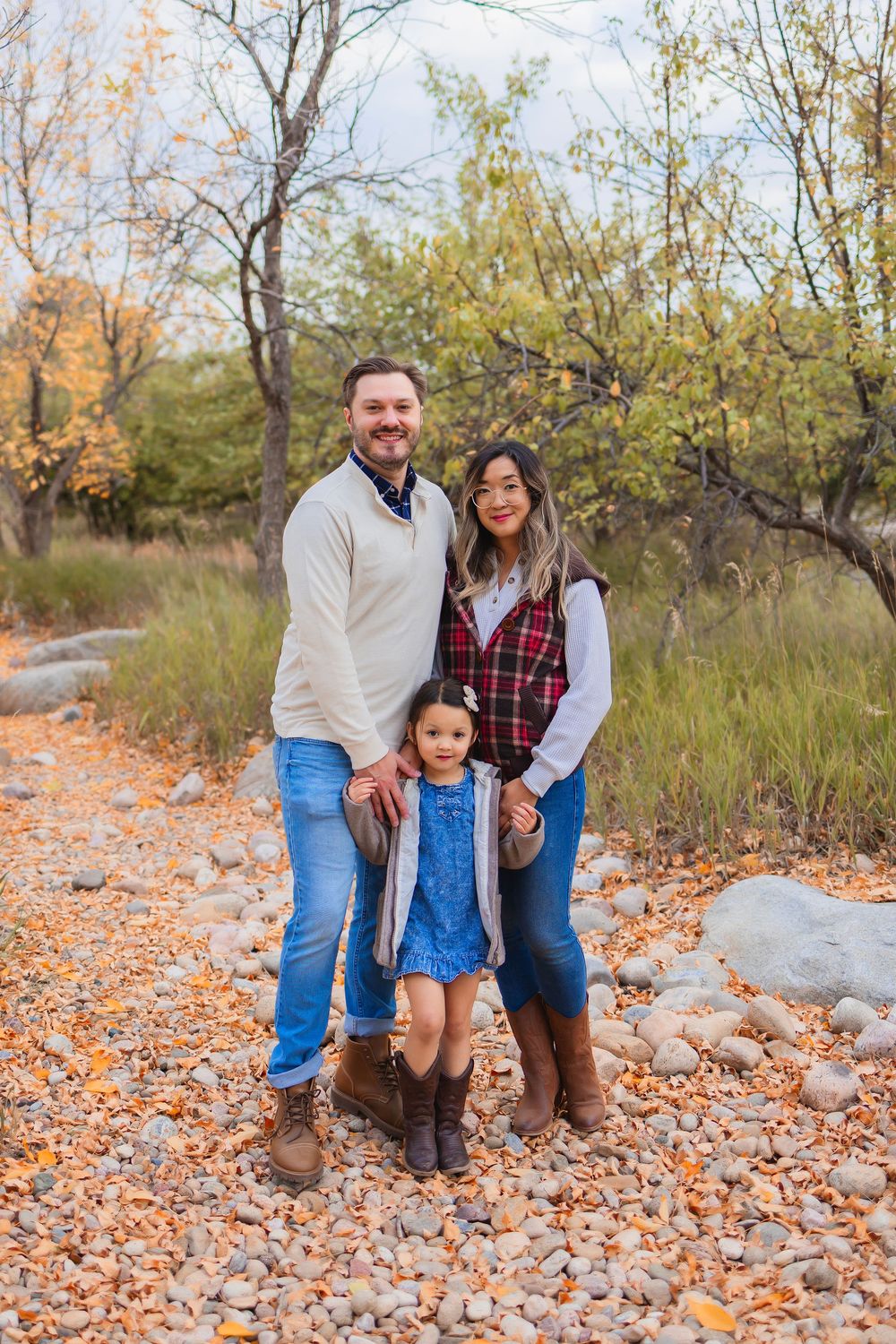 Family of three posing outdoors in fall foliage, parents and young daughter in coordinated outfits on rocky path.