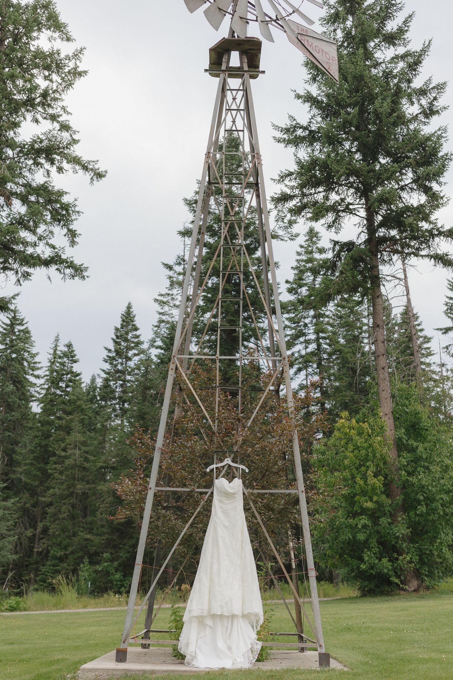 A white wedding dress hangs from a rustic metal windmill structure surrounded by tall evergreen trees.