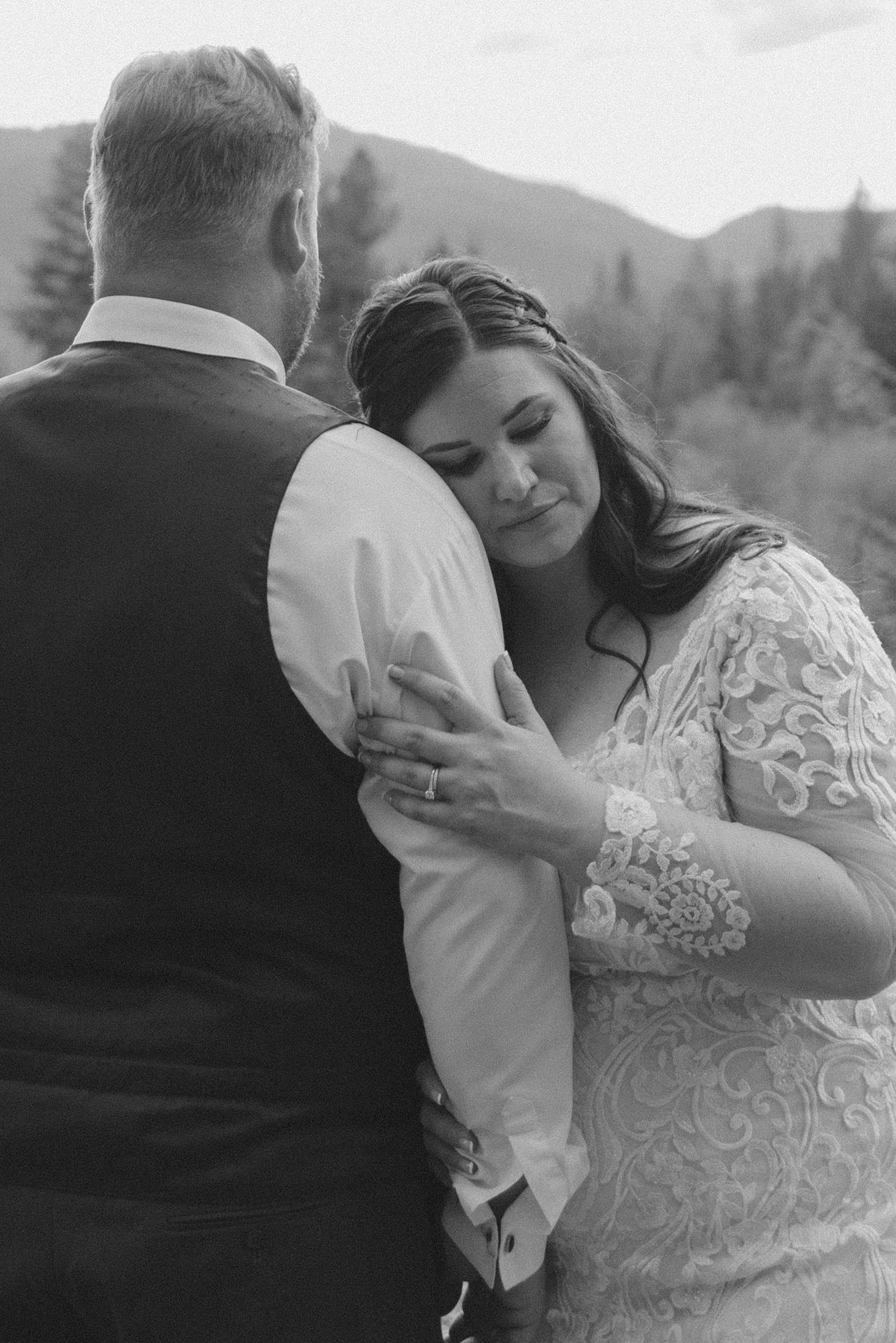 An intimate black and white wedding photo showing a couple embracing outdoors with mountains visible in the background.