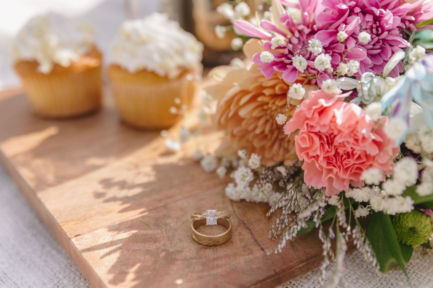 Wedding rings on wooden surface with pink floral bouquet and cupcakes in warm sunlight.