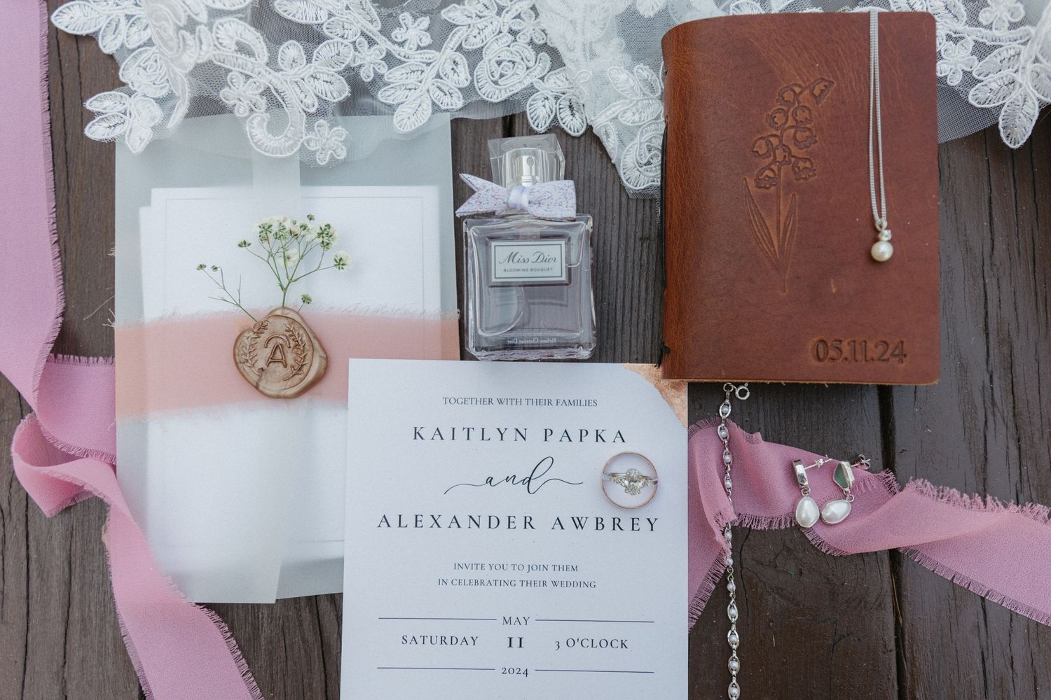 Wedding invitation suite with pink ribbon, perfume bottle, and brown leather journal on wooden surface with lace details.
