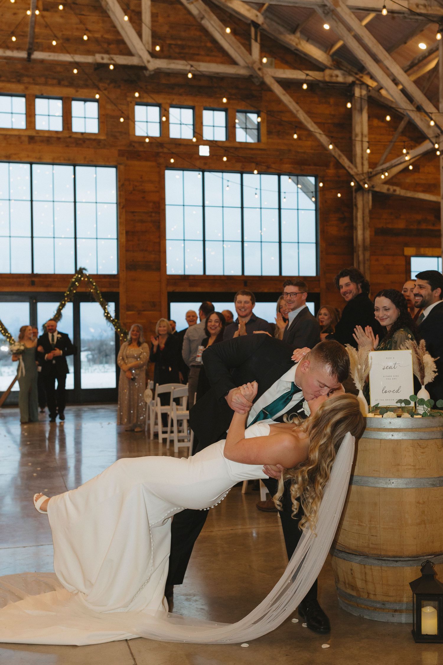 A couple celebrates during their rustic barn wedding reception with glowing string lights and wooden wine barrels visible.