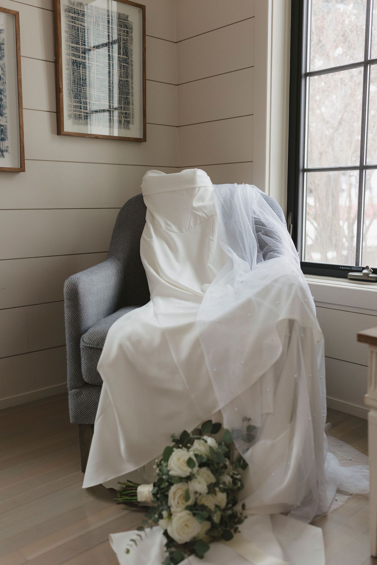 White wedding dress elegantly draped over a gray chair near window with bridal bouquet of white roses on floor.