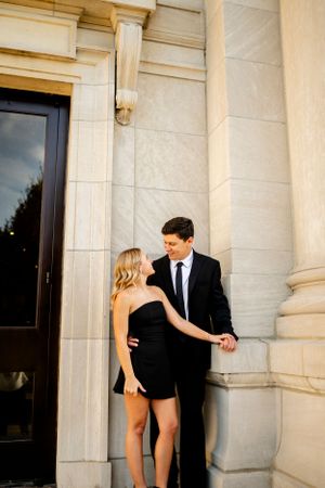 An elegant couple poses against classic white stone columns during a formal photoshoot.