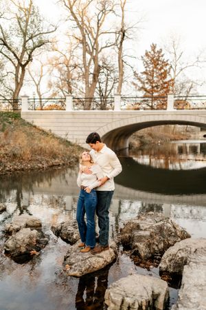 Romantic couple poses by a stone bridge over reflective water during an outdoor photoshoot.
