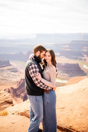 Couple embracing on red rock cliff edge with vast canyon views in background.