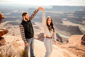 Couple embracing on cliff edge with panoramic desert canyon vista behind them.