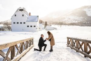 A winter proposal scene on a snow-covered wooden bridge with white buildings in background.
