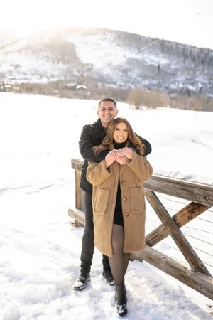 A couple shares joyful moments on a snow-covered bridge with mountain views.