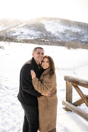 A winter engagement photoshoot with a couple embracing on a snowy bridge.