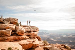 Two people standing on red rock formation with expansive canyon views in background.