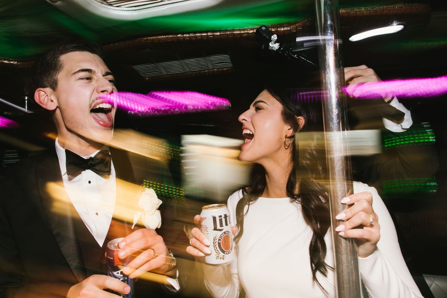 A couple in formal attire laughs enthusiastically while holding drinks in a party bus with colorful lighting.