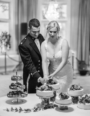 Black and white photo of a couple cutting their wedding cake together at an elegant dessert display with tiered stands.