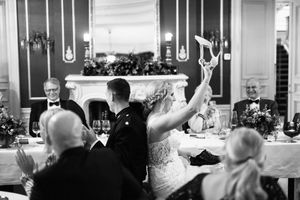 Black and white wedding reception photo showing a high-kicking celebratory moment among guests at a dining table.