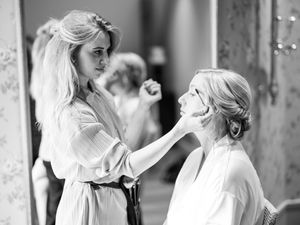 Black and white photo of makeup application in progress with one person doing makeup on another wearing white.